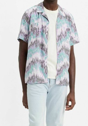 Uomo che indossa una camicia a maniche corte con bottoni e motivo astratto viola, turchese e grigio sopra una t-shirt bianca e jeans azzurri.