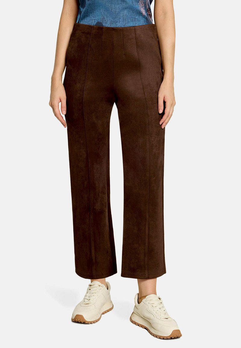 Femme portant un pantalon marron taille haute à jambes larges et des baskets blanches, debout les mains détendues le long du corps sur fond blanc.
