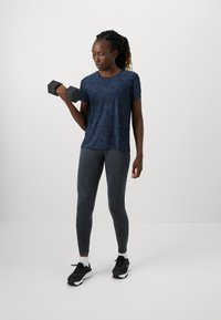 Mørkeblå atletisk t-shirt med et tekstureret mønster, parret med grå leggings, sorte sneakers og en håndvægt holdt i den ene hånd.