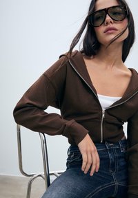 Hnedá skrátená mikina na zips s kapucňou, textúrovaná látka, doladená s tmavomodrými priliehajúcimi džínsami. Modelka má na sebe veľké čierne slnečné okuliare.