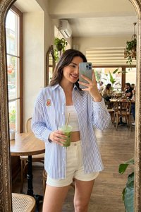 Mladá žena v pruhovanom tričku a bielych šortkách si v kaviarni plnej rastlín a dreveného nábytku robí selfie pred zrkadlom, pričom drží zelené smoothie.