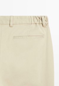 Pantalons beige avec une taille élastique, des passants de ceinture et une poche arrière avec une bordure cousue. Texture de tissu lisse et design ajusté.