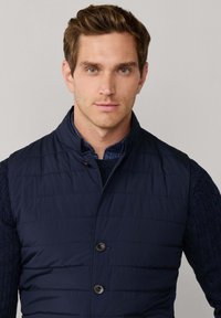 Marineblaue gesteppte Weste mit Stehkragen, ausgestattet mit zwei vorderen Knöpfen und einem darunter liegenden strukturierten marineblauen Strickpullover. Schlichter Hintergrund.