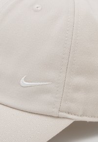 Μπεζ καπέλο από ριγωτό ύφασμα, με καμπυλωτό γείσο και λευκό κεντημένο λογότυπο Nike. Ο σχεδιασμός περιλαμβάνει ραμμένα αξεσουάρ κατά μήκος των ραφών.