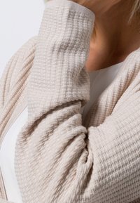 Cardigan beige clair avec un motif texturé en relief. Présente des épaules tombantes et des manches larges, fermeture visible à l'avant.