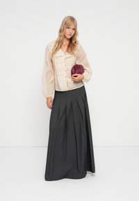 Beige, plisserte bluse med knappelukking og gjennomsiktige ermer, parret med et langt, grått, plisset skjørt. Holder en liten, burgunder veske.