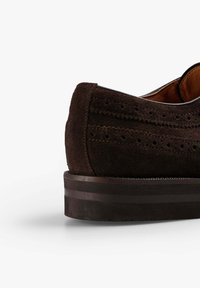 Zapato de ante marrón con trasera redondeada, que presenta detalles de brogue y una suela de goma plana oscura. Textura suave con sutiles acentos de costura.