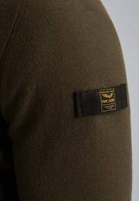 Olivezy zielony materiał o teksturowanym wykończeniu. Posiada czarną, tkana metkę z złotym tekstem i emblematem, przyszytą do rękawa.