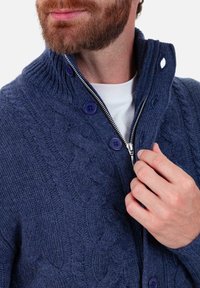 Bärtiger Mann trägt einen marineblauen, kabelgestrickten Pullover mit Knöpfen, teilweise über einem weißen Hemd hochgezogen, und hält am Reißverschluss.