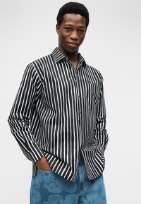 Νεαρός με κοντά σγουρά μαλλιά φοράει μαύρο και άσπρο πουκάμισο με κάθετες ρίγες και μπλε παντελόνι με ανθικό μοτίβο, με τα χέρια στις τσέπες.