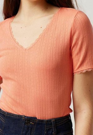 Kvinde iført en koralfarvet tekstureret strikket top med korte ærmer, bølget V-hals og mørkeblå denimjeans.