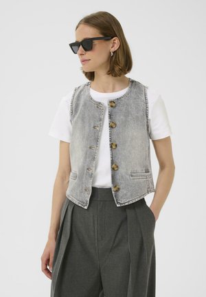 Denim-Weste in Hellgrau mit rundem Ausschnitt und Holzknöpfen. Über einem weißen T-Shirt getragen, kombiniert mit grauen, plissierten Hosen.