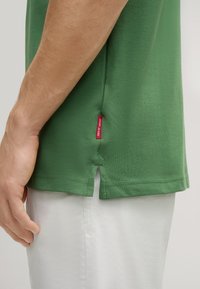 Grünes Kurzarmhemd aus Baumwolle, mit seitlichen Schlitzen am Saum und einem roten Etikett an der linken unteren Naht.