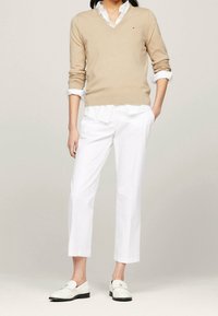 Kobieta ubrana w beżowy sweter z dekoltem w serek na białej koszuli z ko�łnierzykiem, białe spodnie 7/8 i białe loafersy, stojąca z jedną ręką w kieszeni.