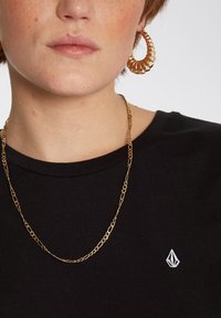 Zwarte T-shirt met een klein wit logo, gecombineerd met een gouden ketting en spiraalvormige gouden ringoorbellen. Soepele stoftextuur.