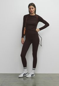 Bordeauxrode top met lange mouwen en leggings van rekbare stof, gecombineerd met witte sneakers en gestreepte sokken, met een kleine bruine tas in de hand.