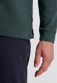 Mannelijke hand ontspannen naast het lichaam, gekleed in donkere broek en een groen visseneffect patroon met lange mouwen, met een Pierre Cardin-label.