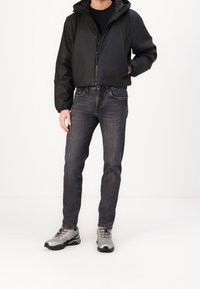 Schwarze Kapuzenjacke mit Reißverschluss, kombiniert mit dunkelgrauen Jeans. Graue Sportschuhe mit schwarzen Akzenten. Lässiges Outfit, präsentiert von einem Model.