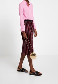 Camicia rosa con bottoni, gonna nera a righe lunga fino al ginocchio, sandali neri con borchie, mentre tiene una borsa rotonda dorata.