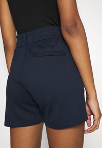 Pantaloni corti blu navy con cintura elastica, dotati di una tasca piatta sul retro e una texture liscia, tagliati sopra la coscia per una vestibilità rilassata.