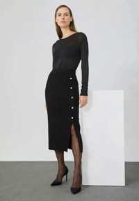 Czarna prążkowana dzianinowa spódnica z rozcięciem po boku, ozdobiona białymi guzikami, zestawiona z dopasowaną czarną bluzką z długim rękawem i czarnymi szpilkami.