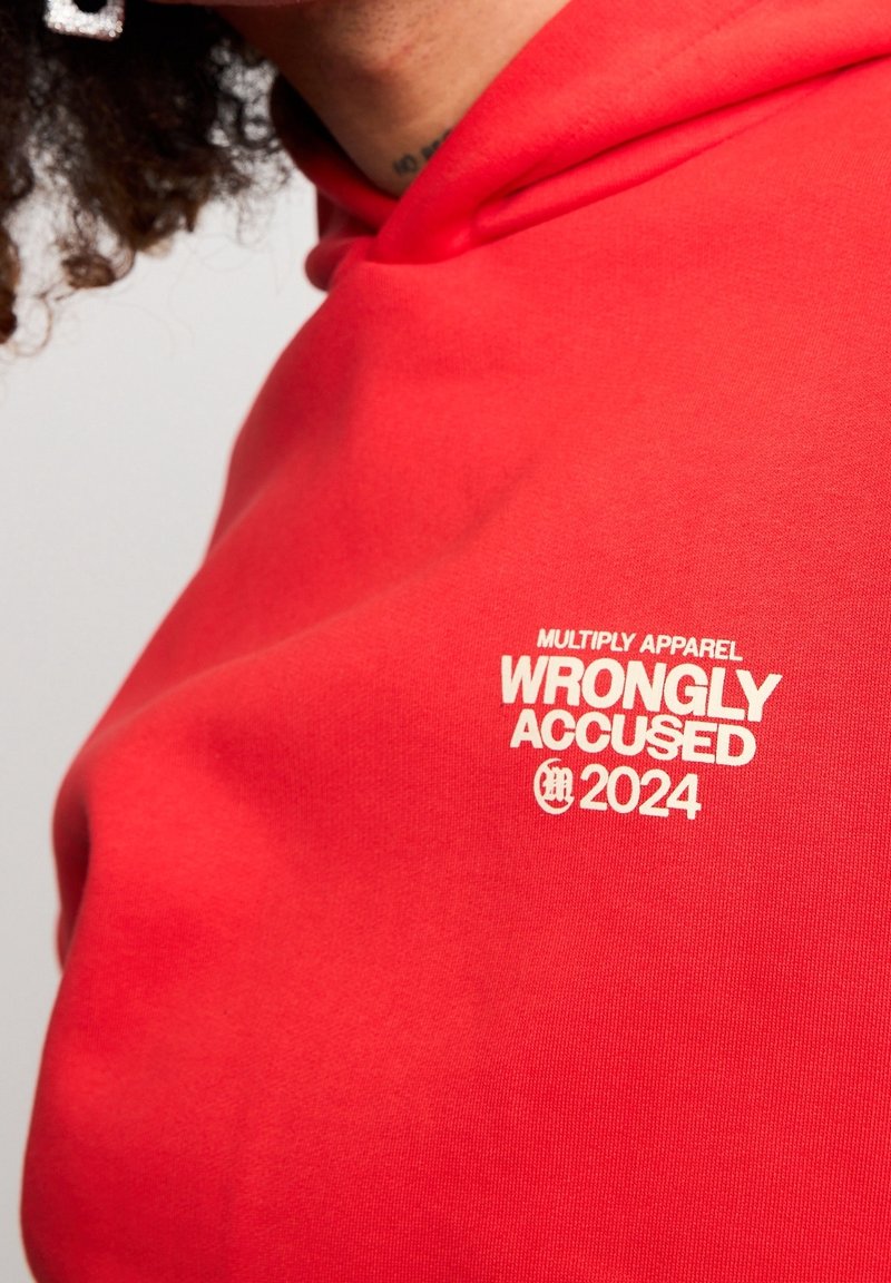 Personne portant un sweat à capuche rouge avec un texte blanc disant "Multiply Apparel Wrongly Accused 2024" près de la poitrine.