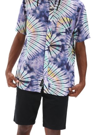 Hombre que lleva una camisa de manga corta con botones y teñida con técnica tie-dye multicolor, y pantalones cortos negros, sujetando el dobladillo de la camisa con ambas manos.