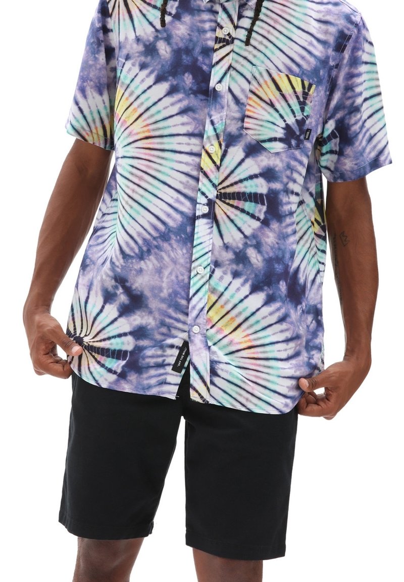 Homme portant une chemise à manches courtes multicolore tie-dye à boutons et un short noir, tenant l'ourlet de la chemise avec les deux mains.