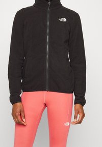 Veste en polaire noire avec col montant et fermeture éclair ; arbore le logo The North Face sur la poitrine gauche ; portée sur desleggings roses.