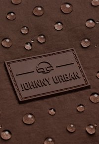 Brun tyg med upphöjda vattendroppar och en texturerad logotypetikett märkt "JOHNNY URBAN". Ytan har en slät finish.