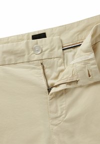 Beige byxor med knäppning och dragkedja, med en slät yta och sydda detaljer vid midjan och ficköppningarna.