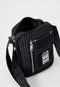 Borsa a tracolla nera in tessuto con righe bianche, chiusura con zip, tasca frontale e patch con il logo Adidas. Include una tracolla regolabile.