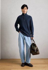 Mørkeblå ribbestrikket sweater med halvlynlås, lyseblå jeans og en grøn taske med brune detaljer på et trægulv.