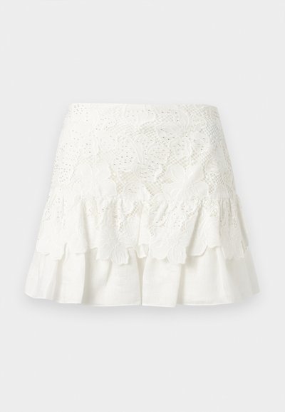 Λευκή μίνι φούστα από δαντέλα με κεντήματα λουλουδιών και βολάν στο τελείωμα, με στρώσεις πάνω σε απλό λευκό ύφασμα.