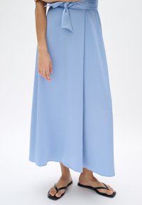 Helle blaue Maxiröcke aus strukturiertem Stoff, mit einem gebundenen Taillendetail, fließendem Design und glattem Saum. Trägt schwarze Flip-Flops.