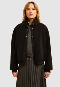 Giacca sherpa nera con alto colletto, chiusura a bottoni a pressione e polsini a costine. Abbinata a una gonna grigia a pieghe e una maglia strutturata.