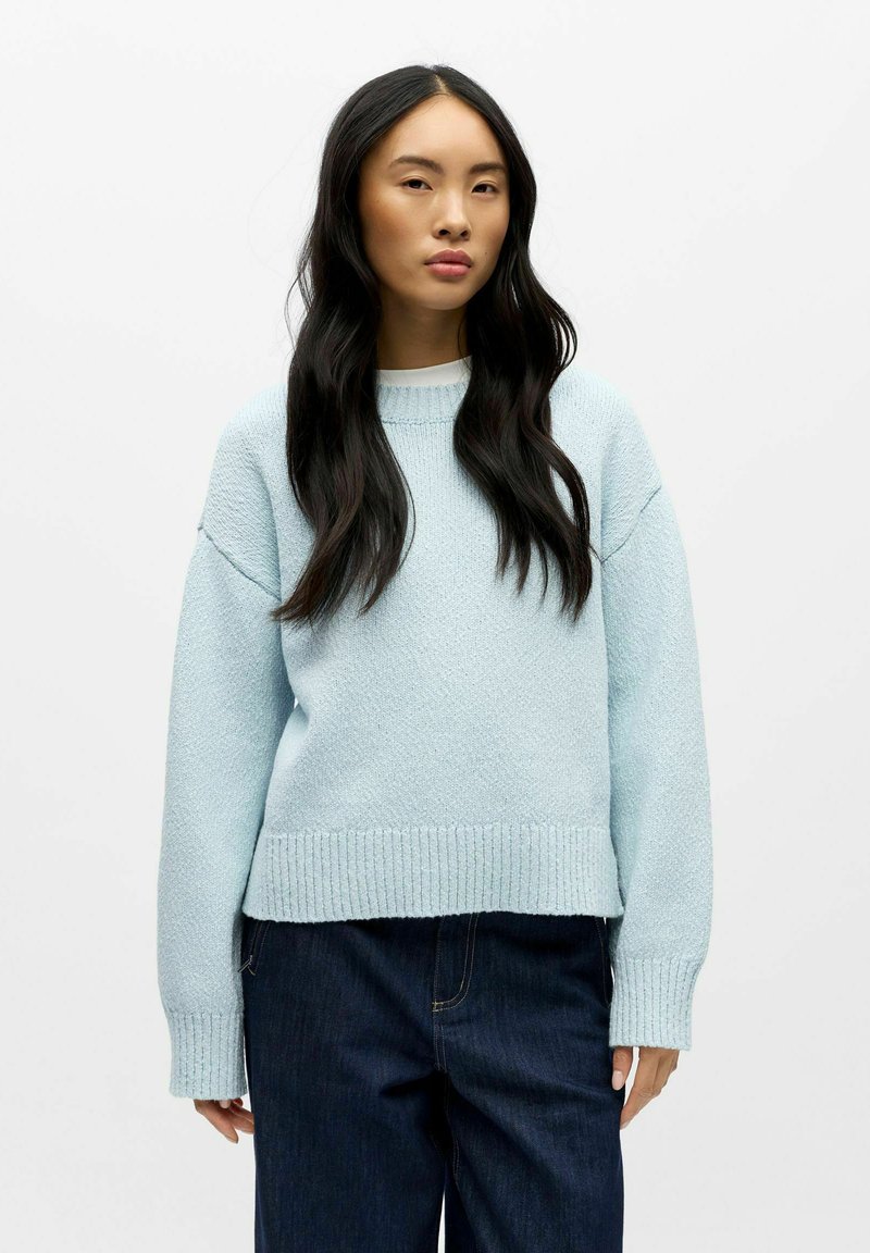 Lys blå strikkegenser med strukturert finish, rund halskant og ribbede mansjetter og kant. Kombinert med mørke denimbukser.