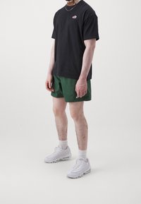 Svart bomullströja med en liten röd logotyp, gröna shorts, vita sneakers och vita ankelsockor på en stående figur mot en enfärgad bakgrund.