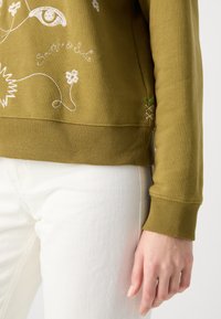 Sweat-shirt vert olive avec broderie florale blanche et design de marque. Présente des poignets et un ourlet côtelés, avec une couture décorative sur le côté.