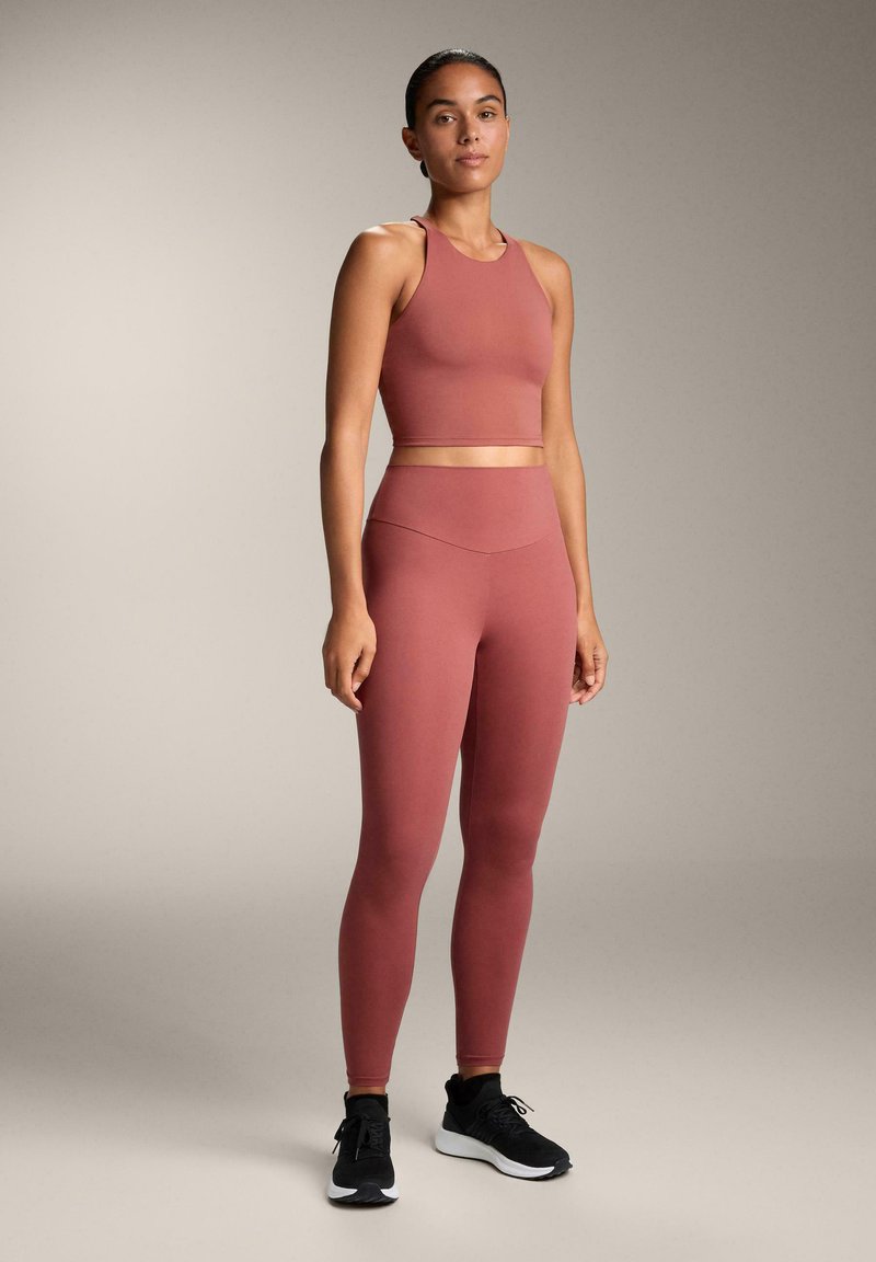 Sutien sport de culoare roz prăfuit și colanți cu talie înaltă din material elastic, asortați cu pantofi sport negri, pe un fundal neutru.