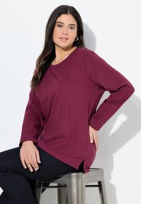 Bordeaux langslavige shirt met een ronde hals, relaxte pasvorm, zijsplitten en gladde stof, gecombineerd met donkere jeans, zittend op een metalen kruk.