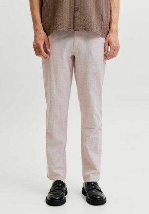 Uomo che indossa pantaloni beige, camicia marrone testurizzata e mocassini in pelle nera, in piedi davanti a uno sfondo chiaro e uniforme.