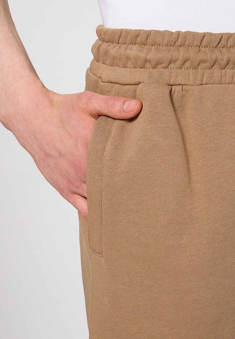 Pantaloni della tuta beige con vita elastica, tasche laterali e un morbido tessuto testurizzato. Primo piano di una mano poggiata sulla tasca.
