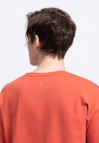 Sweatshirt corail orange avec une coupe décontractée. Présente un petit logo de ciseaux brodé dans le haut du dos. Fabriqué à partir d'un tissu doux et texturé.