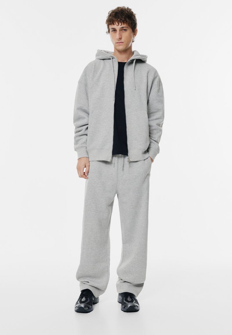 Harmaa fleece-huppari, jossa on kiristysnauhat, yhdistettynä samanvärisiin harmaisiin collegehousuihin. Mallilla on musta T-paita alla ja mustat lenkkarit.