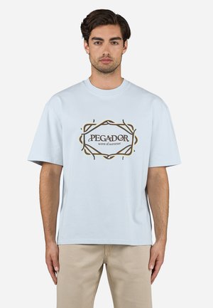 Light blue cotton t-shirt with short sleeves, featuring a graphic print "PEGADOR scent of summer" in dark brown and gold outline.