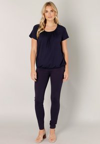 Donkerblauw shirt met korte mouwen en een geplooide halslijn, gecombineerd met nauwsluitende donkerblauwe leggings. Draagt stijgriemhakken. Subtiele textuur.