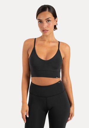 Kvinde iført en sort cropped sportstop med tynde stropper og sorte leggings med høj talje, stående mod en ensfarvet lys baggrund.