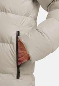 Lichtgrijze pufferjack met een ritszak aan de linkerkant. Gevoerde textuur met gestikte secties, voorzien van een zwarte ritsdetail.