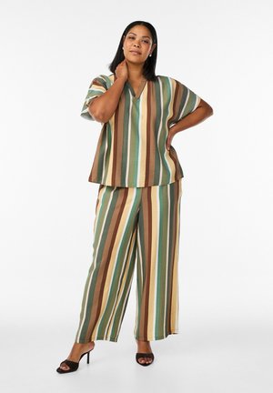 Kvinde iført et matchende sæt med kortærmet top og bukser med vide ben i grønne, brune, beige og blå nuancer, stående med den ene hånd på nakken.